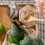 A metal basket with fresh vegetables, avocado, lime, and oats arranged in a flat lay style.