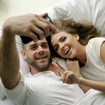 Young beautiful and loving couple take selfie picture on smartphone camera while lying in bed at morning
